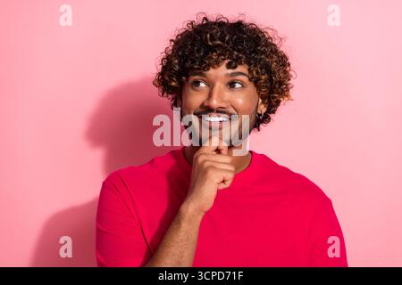 Smiling young man with curly hair in a red shirt on pink background showing thoughtful pose with hand on chin Stock Photo