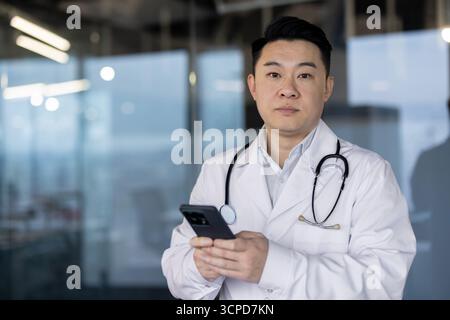 Portrait of a young serious male doctor in a white coat standing in a clinic and using a mobile phone, looking at the camera. Stock Photo