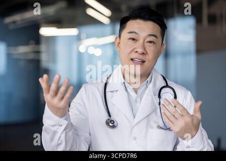 A young Asian doctor stands in the hospital lobby in uniform, talking to the camera and gesturing with his hands. Stock Photo
