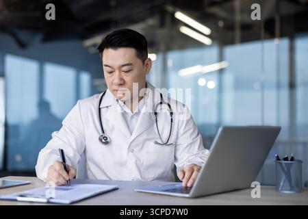 An asian doctor is focused at his desk, writing on documents while using his laptop for work. He looks concentrated. Stock Photo