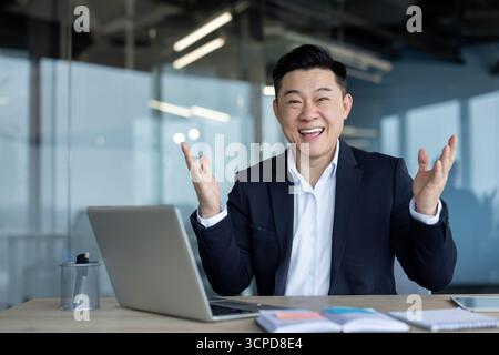 Portrait of a happy and smiling young Asian businessman sitting in the office at a table with a laptop and looking at the camera with satisfaction, gesturing with his hands. Stock Photo