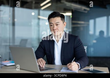 Serious and focused young Asian businessman working in the office on a laptop, taking notes. Stock Photo