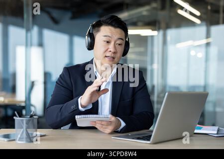 Asian young serious man in headphones and suit sitting in the office in front of a laptop, talking on a video call, taking notes. Stock Photo