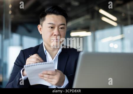 Close-up photo of a young concentrated Asian man in a suit sitting in an office, looking at a laptop screen and making notes in a notebook. Stock Photo