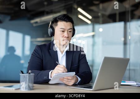 A thoughtful and focused young man wearing headphones sits at a desk in the office, listening to an online lecture meeting and taking notes in a notebook. Stock Photo