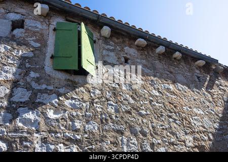 An inviting rustic scene featuring an open green shuttered window set in a rough stone wall beneath a bright blue sky. Perfect for architecture, rural Stock Photo