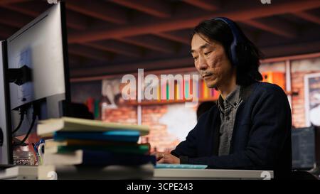 Senior admin coding advanced AI algorithm in startup office, refining machine learning models to enhance automation, wearing headphones. CTO manager listening music and coding, camera B Stock Photo