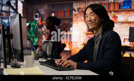 Portrait of smiling senior engineer coding advanced AI algorithm in startup office, refining machine learning models. Cheerful man using coding platform in Silicon Valley workplace, camera B Stock Photo