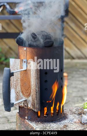 A metal charcoal chimney starter sits on a stone surface, filled with charcoal briquettes, flames rising and smoke billowing from the top. Stock Photo