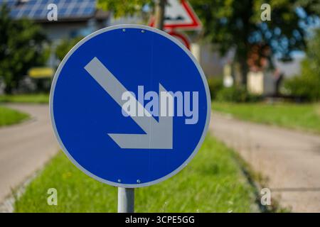 A close-up shows a blue and grey road sign with a directional arrow pointing downwards and to the left, against a blurred background. Stock Photo