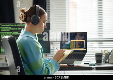 Young Woman Wearing Headphones Writing Notes While Coding at Desk Stock Photo