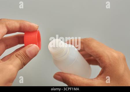 hands opening or unscrewing red ribbed cap on small translucent white plastic bottle, access to medicine or remedy, isolated gray background Stock Photo