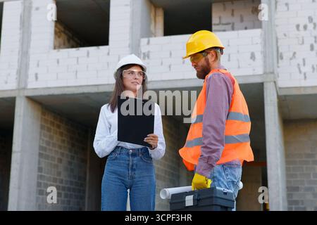 Female architect holding clipboard and male construction worker carrying blueprints and toolbox are inspecting building progress Stock Photo