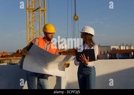Construction workers reviewing blueprints on a building site with crane in the background, discussing project plans Stock Photo