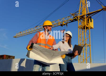 Construction workers reviewing blueprints on a building site with crane in the background, discussing project plans Stock Photo
