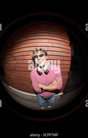 Fish eye portrait of young Caucasian man smiling at camera wearing headphones around neck and sunglasses, standing outdoors against brick wall with arms crossed Stock Photo
