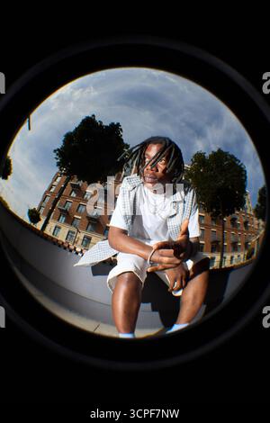 Fish eye view of Black teenager boy sitting outdoors in urban setting, looking away from camera with thoughtful expression, wearing casual clothing, surrounded by buildings and trees Stock Photo