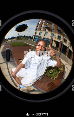 Fish eye portrait of Black teenager boy sitting on outdoor bench making hand gesture and smiling at camera, urban background with modern building and greenery visible in scene Stock Photo