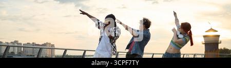 Three teenagers, including Black, Caucasian boy and girl friends, standing outdoors on urban waterfront, performing dab dance pose together with city skyline in background Stock Photo