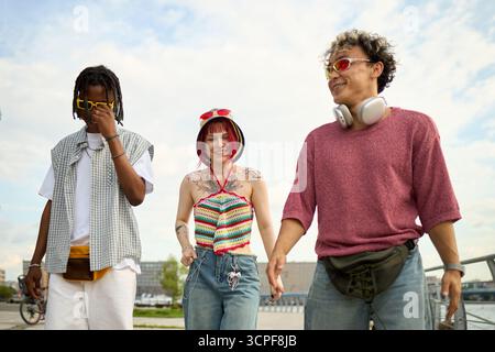 Group of three young people, multiethnic men and Caucasian woman, walking outdoors together, smiling and interacting, wearing sunglasses and headphones, enjoying urban environment Stock Photo