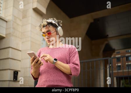 Young Asian man wearing headphones and sunglasses using smartphone outdoors, standing in urban setting with focused expression, interacting with device Stock Photo