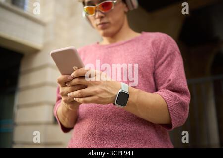 Young Caucasian man wearing headphones and sunglasses using smartphone outdoors, holding device with both hands, smartwatch visible on wrist, standing near building Stock Photo