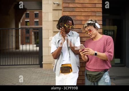 Young Black man and Caucasian friend standing outdoors, smiling and looking at smartphone together, wearing sunglasses and headphones, enjoying leisure time in urban setting Stock Photo