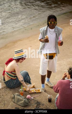 Young Caucasian woman with red hair sitting on ground eating fast food with drink, Black man standing holding sandwich and beverage, young male friend sitting nearby Stock Photo