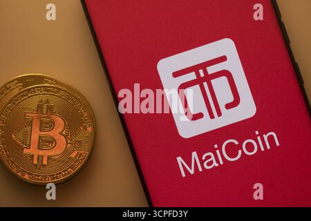 Bitcoin coin next to a smartphone featuring the MaiCoin logo on a red background Stock Photo