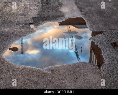 Puddle on rough asphalt reflects clouds lit by sunrise, along with a streetlight and construction cranes Stock Photo