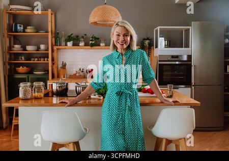Happy mature woman standing in her modern kitchen with various fresh vegetables on the counter, ready to prepare a healthy meal, promoting a wellness Stock Photo
