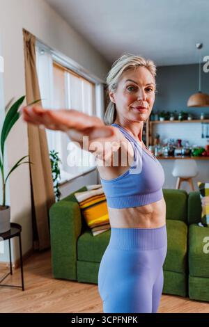 Fit mature woman practicing yoga, enjoying healthy lifestyle and exercising in her living room, holding warrior two pose with arms outstretched and fo Stock Photo