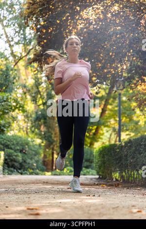Young blonde woman running along a park path, focused on fitness and outdoor training, jogging in nature for health, endurance and active lifestyle en Stock Photo