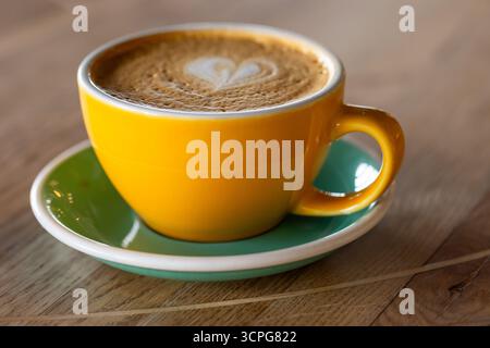 Cappuccino. A vivid yellow coffee cup sits on a green saucer, filled with frothy latte featuring a heart-shaped art. Perfect for morning rituals, cafe Stock Photo
