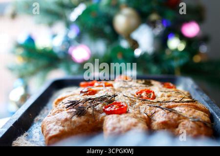 Fresh homemade focaccia bread on a baking tray in front of a decorated Christmas tree with colorful ornaments Stock Photo