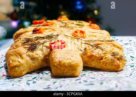 Homemade tree-shaped focaccia bread with cherry tomatoes and rosemary on floral tablecloth near a decorated Christmas tree Stock Photo