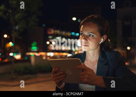 Female using digital tablet with earbuds outdoors in urban evening Stock Photo