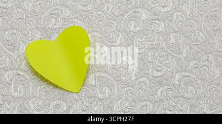 Delicate contrast. Yellow heart-shaped note lies gently on a textured, floral-patterned white fabric, creating a contrast between the smooth paper and Stock Photo