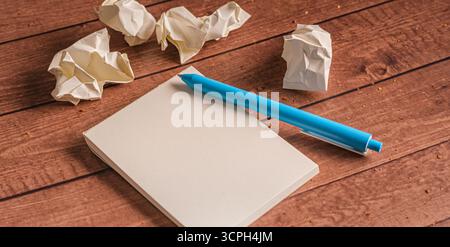 A blue pen rests beside a blank notepad, surrounded by crumpled paper balls on a wooden surface. Capturing the essence of creativity and the process o Stock Photo
