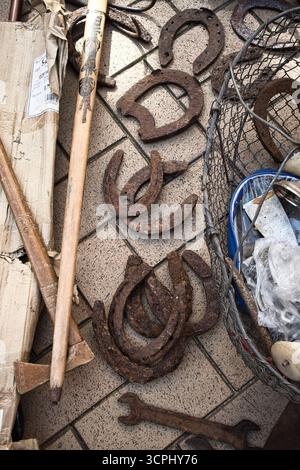 A pile of rusty horseshoes and a wooden stick. The horseshoes are scattered on the ground and the stick is leaning against a wall. The scene gives off Stock Photo