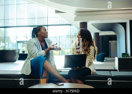 Two female coworkers are engaged in a lively discussion in a modern office setting. They are seated on a bench, focusing on a laptop, indicating a col Stock Photo