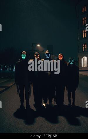 A group of people wearing glowing illuminated masks stands on a dark city street at night, featuring colorful lights and shadows for an eerie and myst Stock Photo