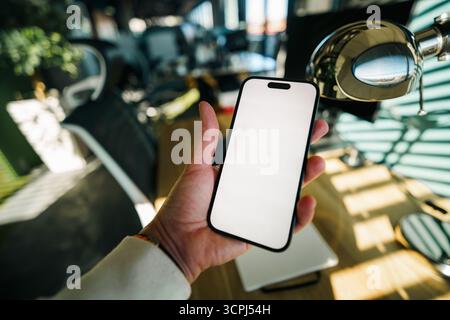 Smartphone mockup office workspace hand holding device. Close-up of a person's hand holding a modern smartphone with blank white screen in a contempor Stock Photo