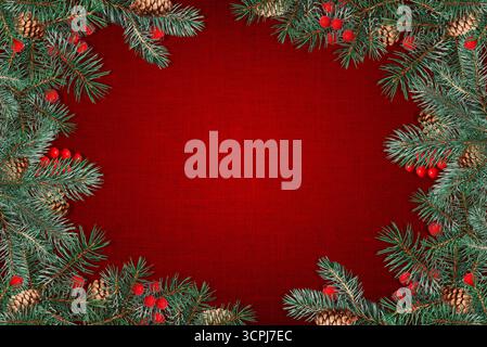 Holiday background with a festive frame of pine branches, cones, and red berries on red canvas texture. Stock Photo