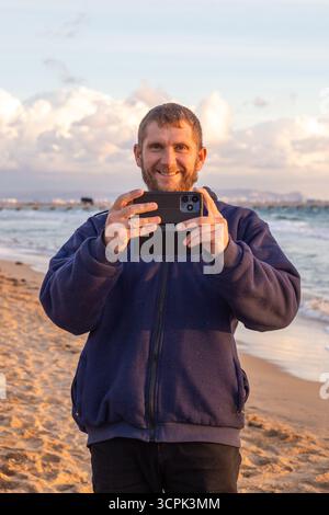 A man stands on a sandy beach and smiles while taking a photo of a picturesque ocean view on his phone under a bright cloudy sky. Stock Photo