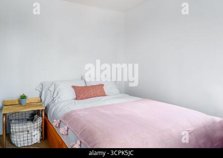 White bedroom with pink chenille pillow, throw blanket, and side table with basket. Side angle view. Stock Photo