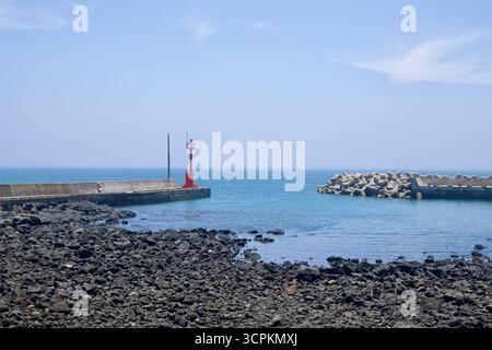 A slim red harbor beacon marks the end of a concrete jetty as tetrapod breakwaters guard the channel, with black basalt rocks exposed at low tide on J Stock Photo