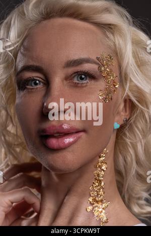 blonde girl smiles confidently showcasing gold glitter adorning her face. She poses gracefully highlighting her unique style and joyful spirit during an artistic moment. Stock Photo