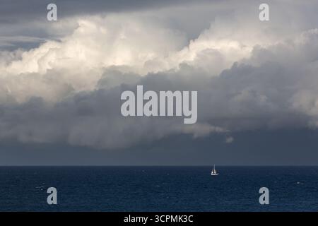 A solitary yacht sails on the open ocean, dwarfed by the vast, turbulent dark blue sea beneath a dramatic sky filled with thick, billowing clouds. Stock Photo