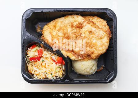 Takeaway schnitzel with potatoes in black plastic tray. Isolated on white background. Fast food lunch box with fried cutlet and vegetables. Stock Photo
